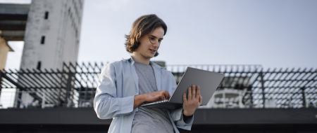 Mężczyzna stojący na zewnątrz trzyma w&nbsp;jednej ręce otwarty laptop Dell Pro Precision&nbsp;14 z&nbsp;serii 7, a&nbsp;drugą ręką na nim pisze.