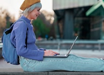 Zdjęcie siedzącej kobiety w fioletowym swetrze, z plecakiem oraz z notebookiem Dell XPS 13 9315, z którego korzysta, na kolanach.
