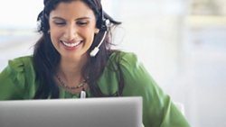 Uśmiechnięta kobieta w zielonej bluzce z zestawem słuchawkowym na głowie korzystająca z notebooka Dell.